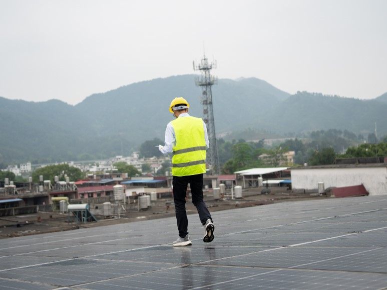 Person in yellow safety vest and hard hat inspecting solar panels on rooftop with mountain backdrop.