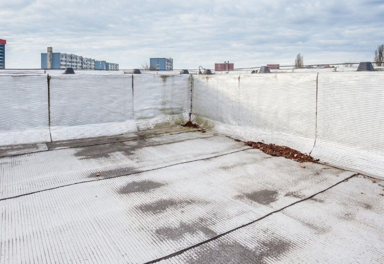 Flat, white commercial rooftop with dark stains and a low barrier fence, cloudy sky, buildings in the distance.