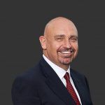 Bald man in suit smiling, looking at the camera. Red tie, dark suit jacket, white shirt. Dark background.