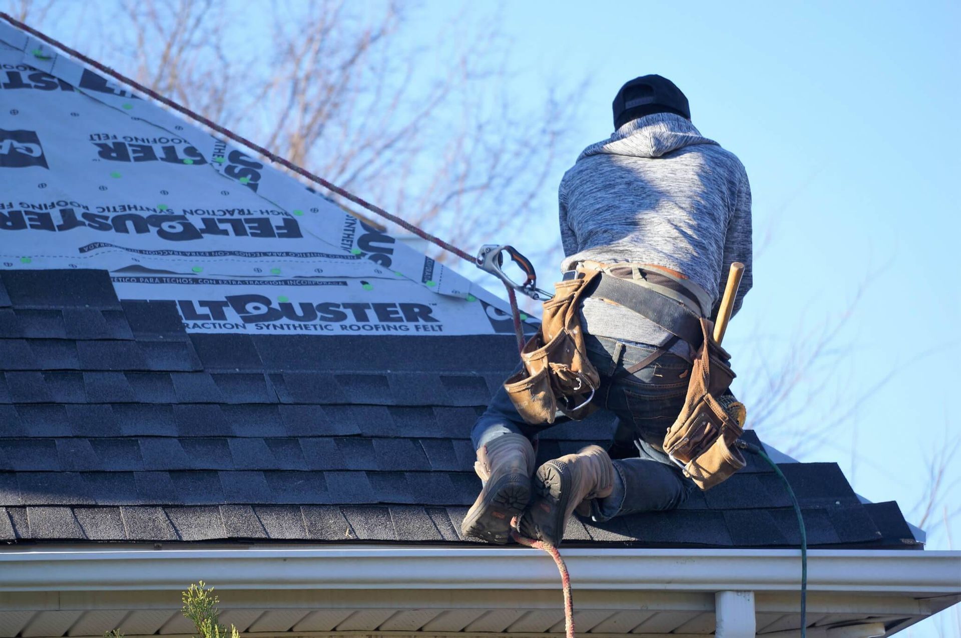 Roofer on a rooftop, laying down new shingles. Blue sky in the background, tool belt visible, working on a home.