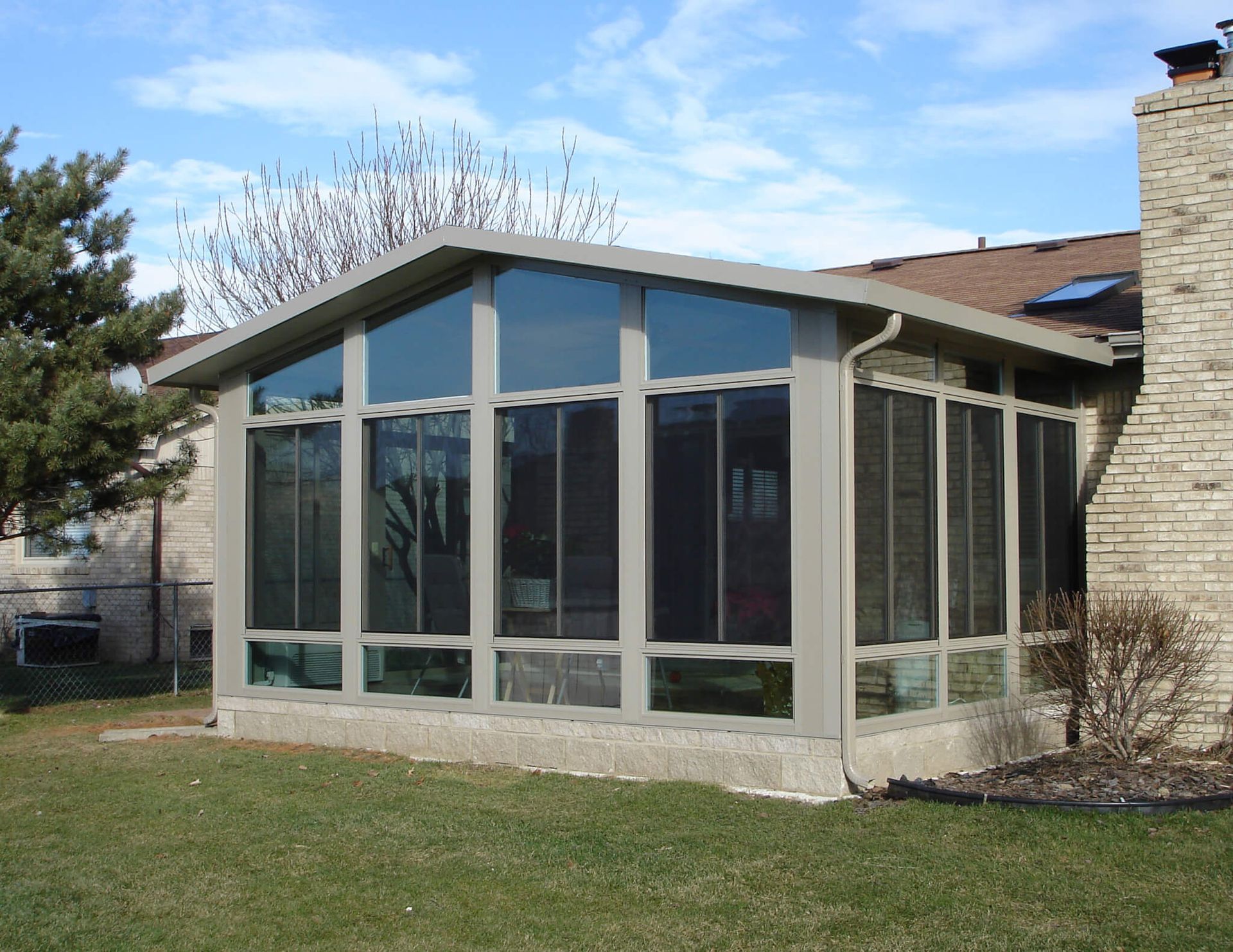 Sunroom addition with large windows and neutral siding, attached to a brick house.