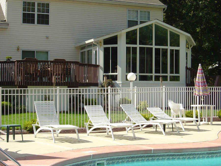 Backyard with pool, deck, and sunroom. White fence, lawn, lounge chairs, and an umbrella near the pool.