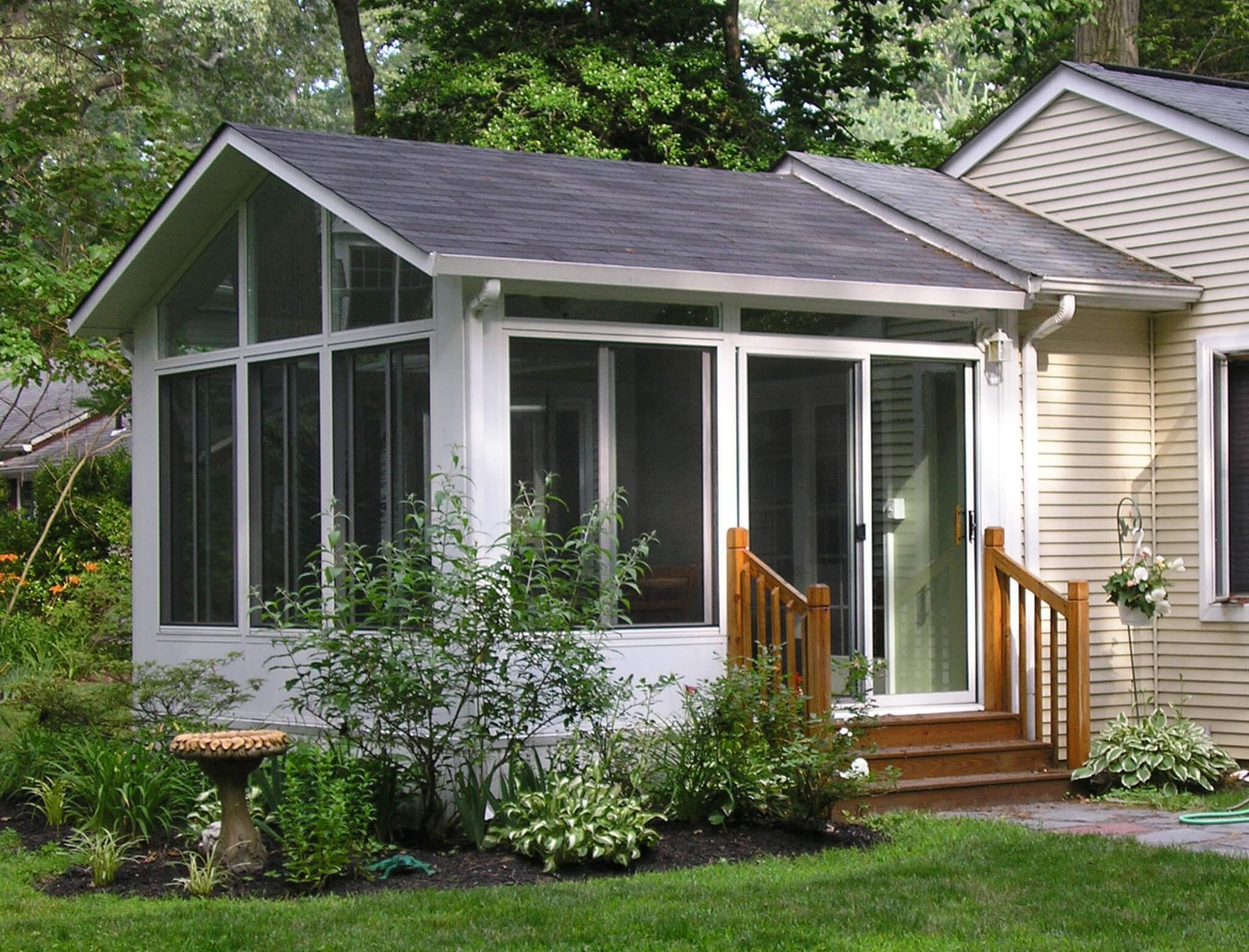 Screened porch addition with white frame, glass sliding door, and wooden steps. Situated next to a house with dark roof and landscaping.