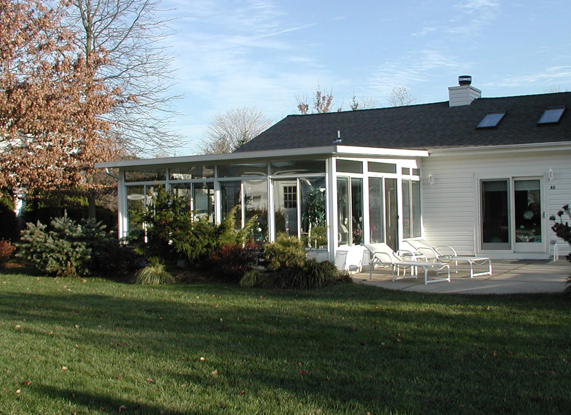 Sunroom addition to a white house with lawn, trees, and patio furniture.