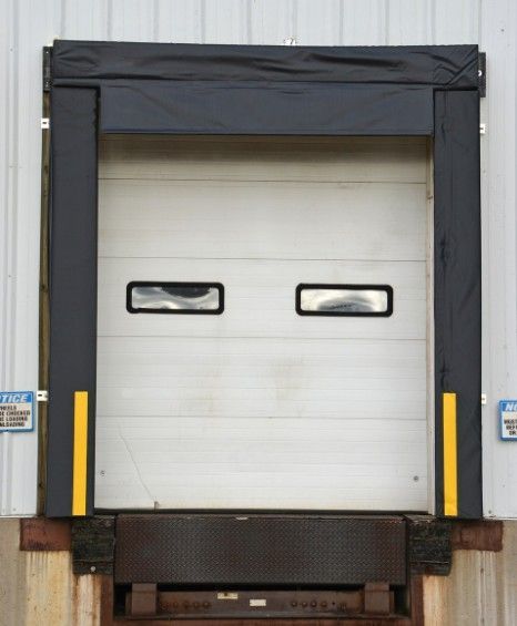 Closed white loading dock door with small windows, framed by black dock seal and yellow safety posts.