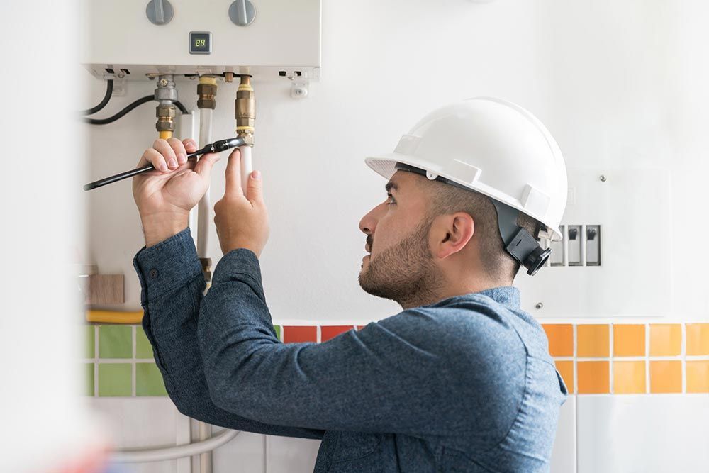 A person in a hard hat repairs plumbing with a wrench, likely in a kitchen or utility room.