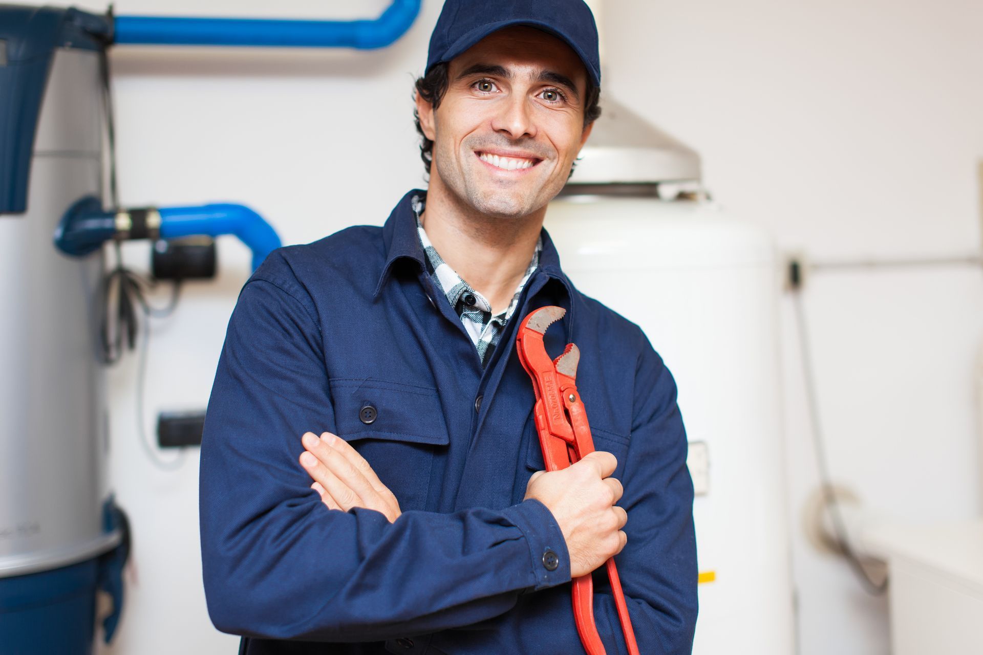 Plumber in blue uniform, holding red wrench, smiling in front of pipes and water heater.