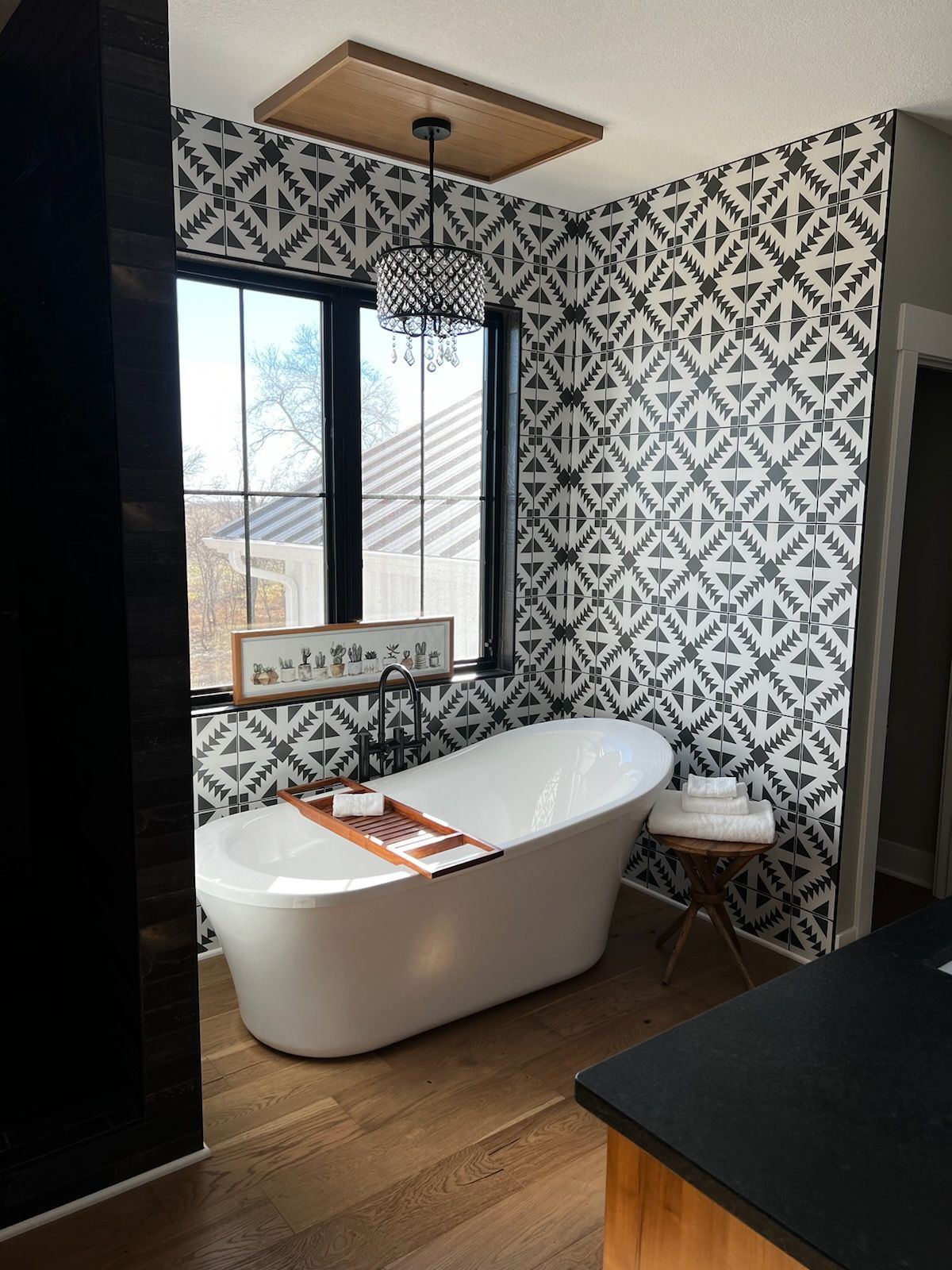 Bathroom with white tub, black and white patterned wall, window with black trim, and wooden floor.