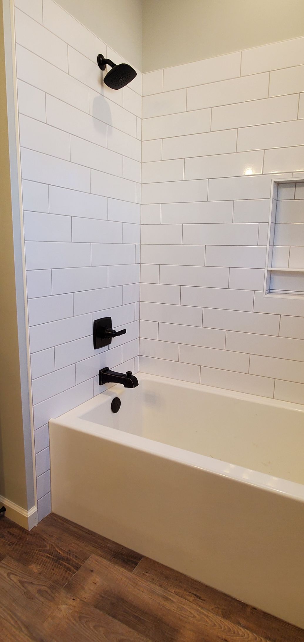 Bathroom with white tile shower, black fixtures, and a white bathtub. Brown wood-look floor.