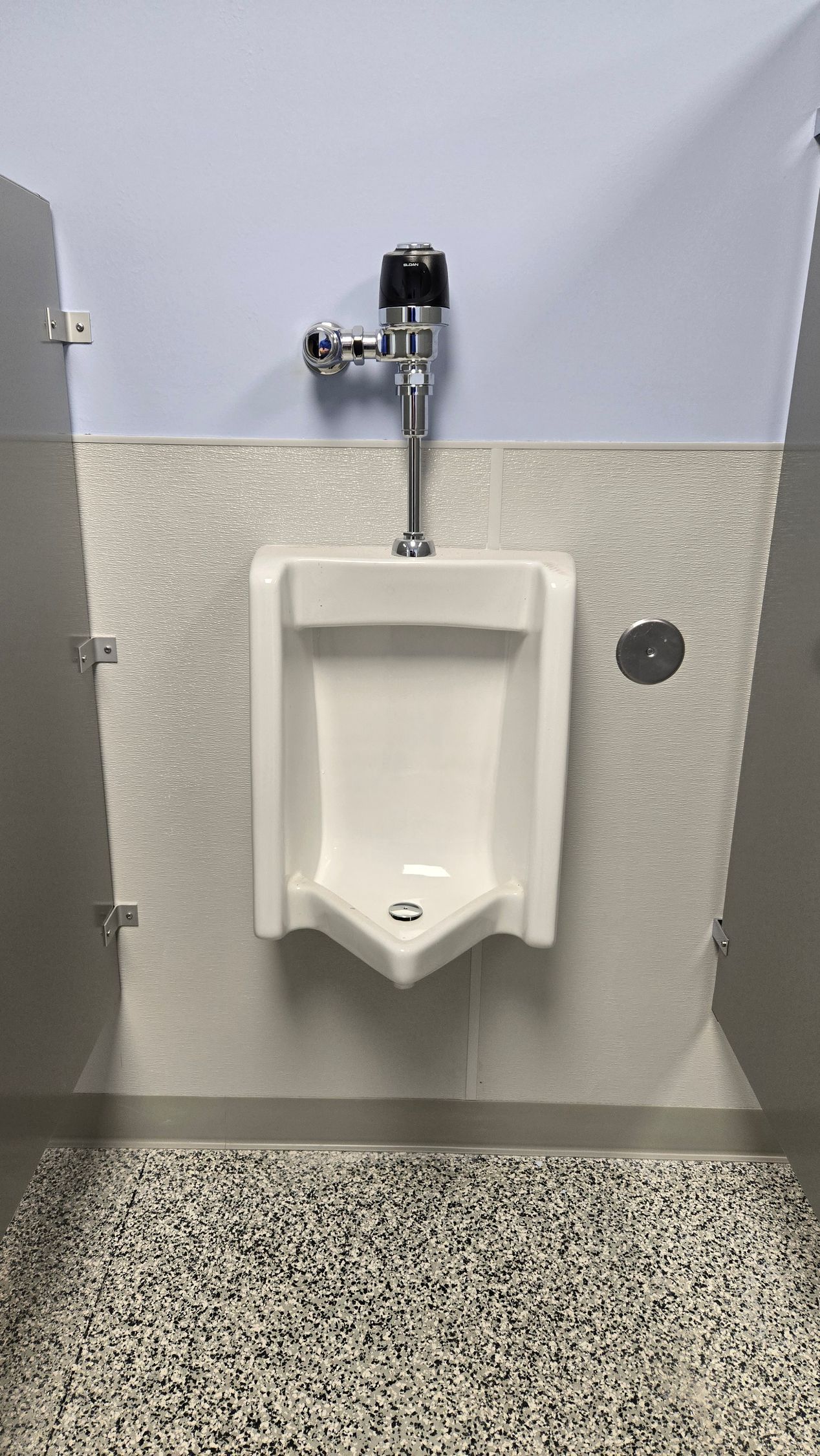 Urinal in a public restroom with grey and white speckled floor and pale blue wall above textured wall tile.