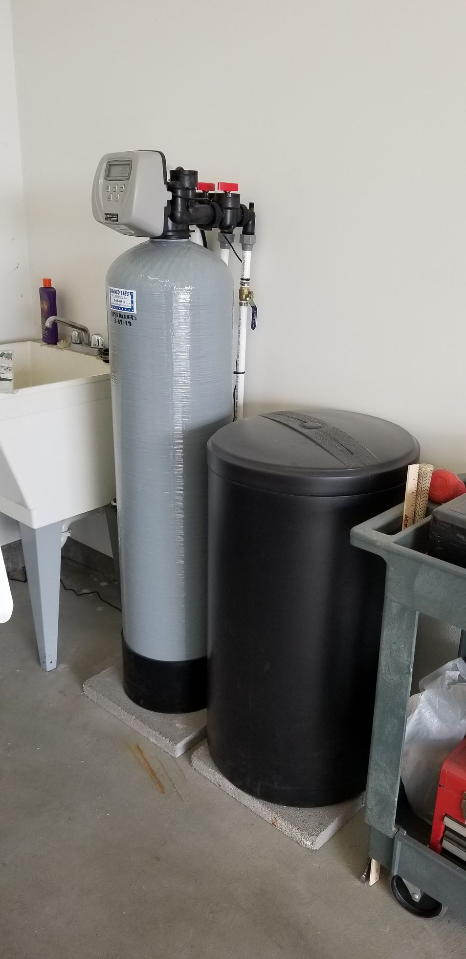 Water softener system in a garage. Tall grey tank and black brine tank next to each other on concrete pads.