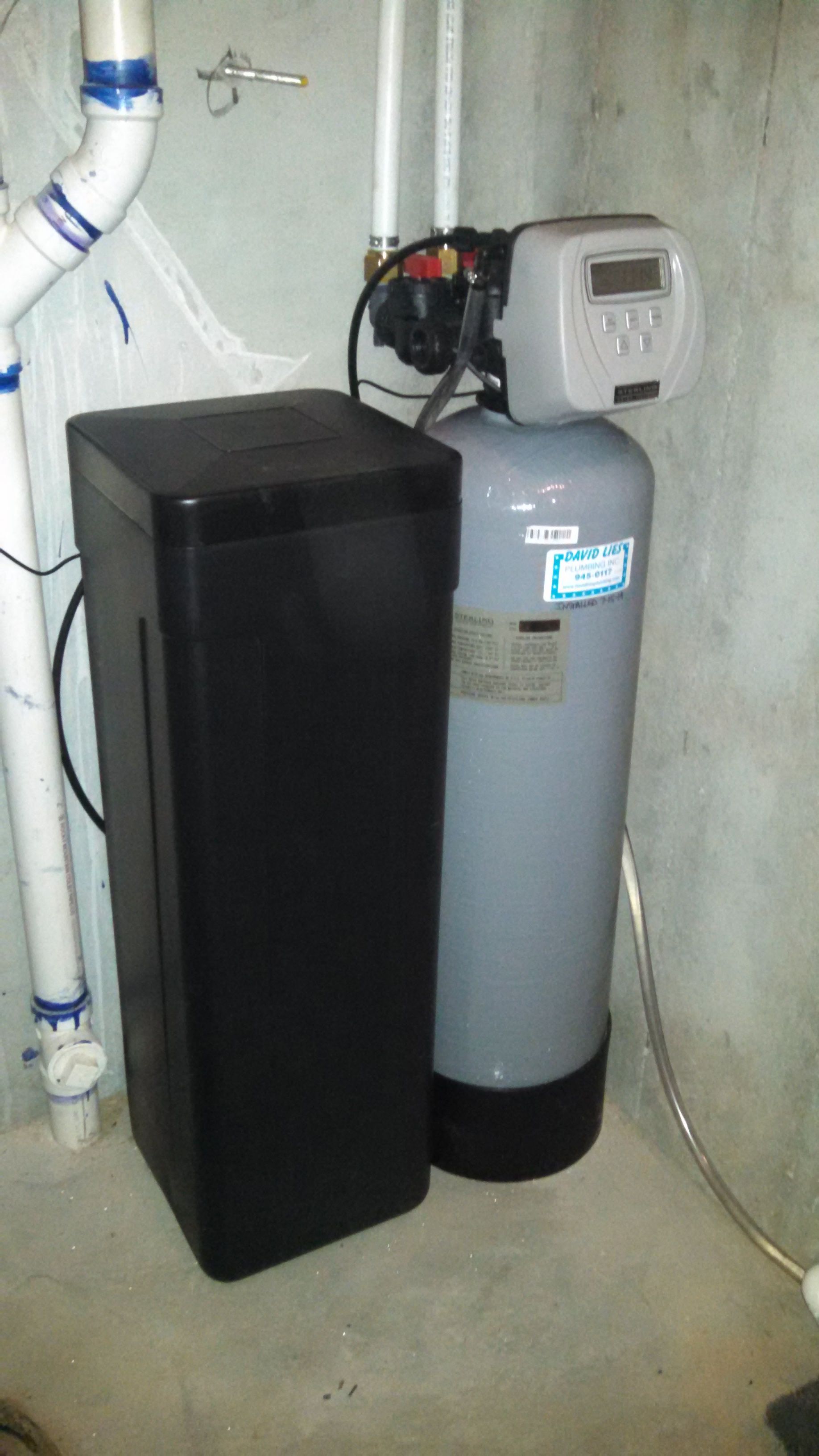 Water softener system in a basement setting; black tank, gray tank with digital display, white pipes.