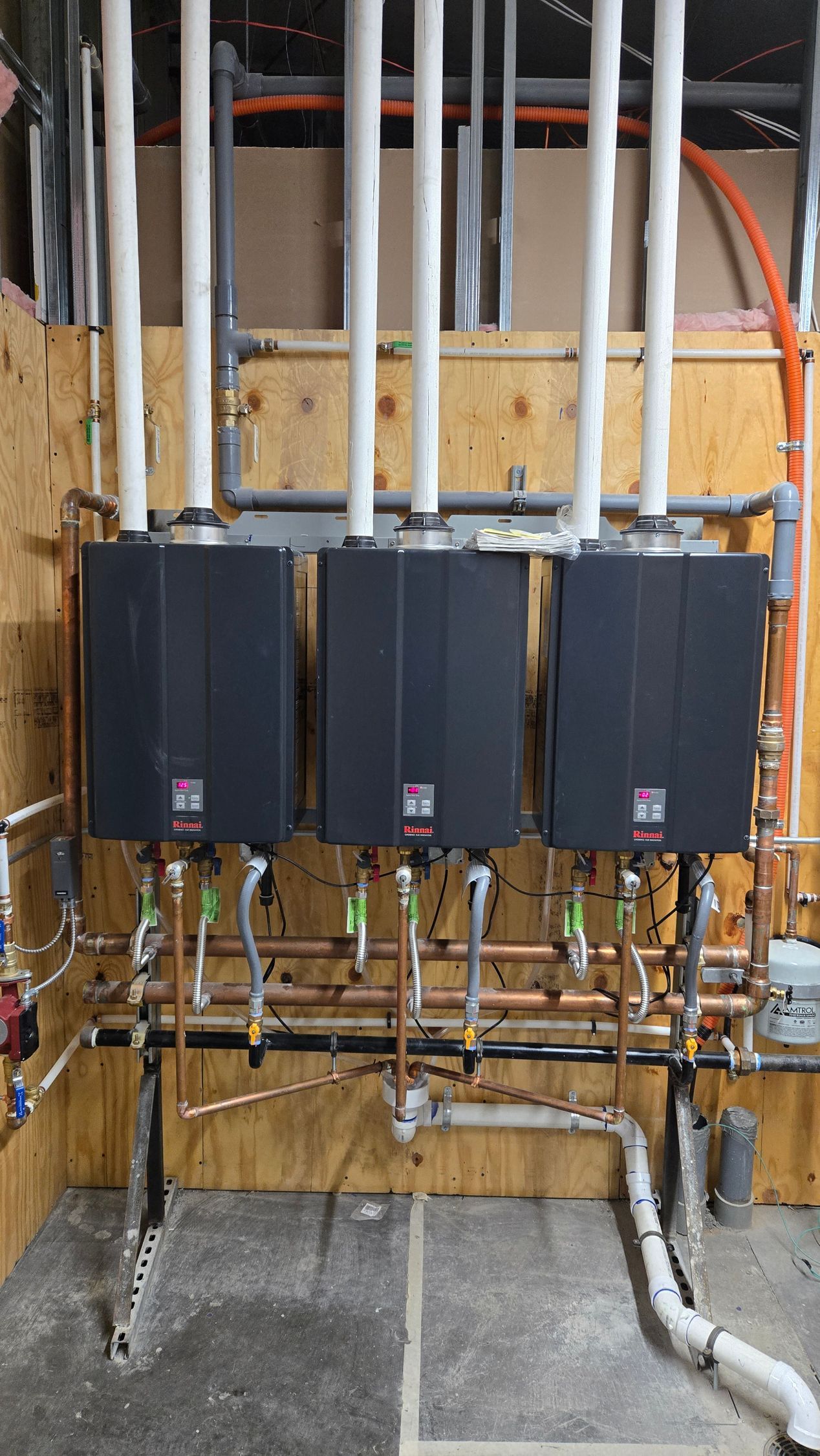 Three black tankless water heaters mounted on a wooden wall with copper and white PVC pipes.