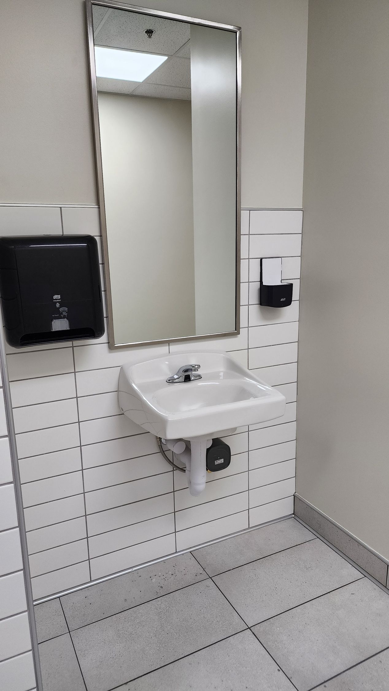 Bathroom with a white sink, mirror, paper towel dispenser, and black soap dispenser on a tiled wall.