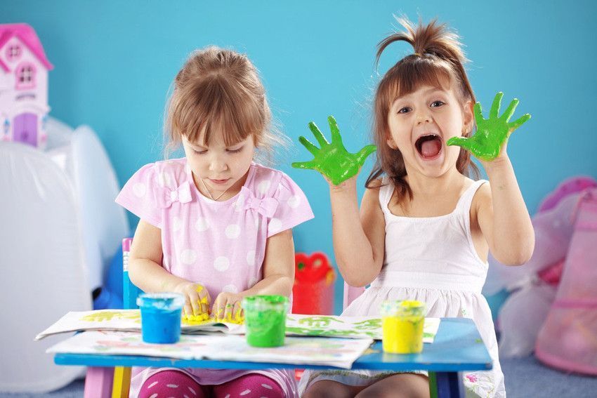 Two children painting at a table; one with green handprints, excited expression, blue background.