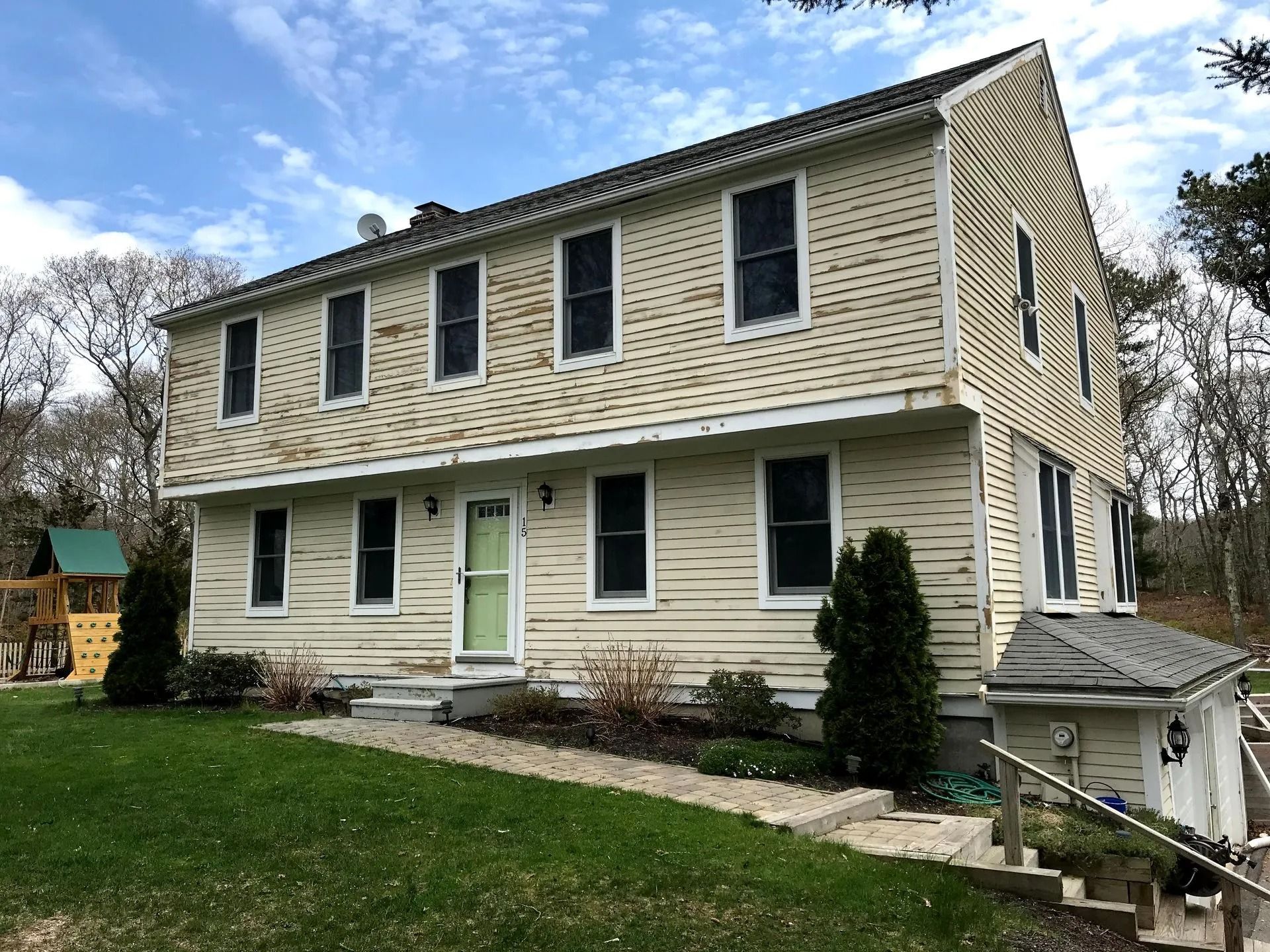 Two-story beige house with multiple windows, green lawn, and a play structure in the yard.