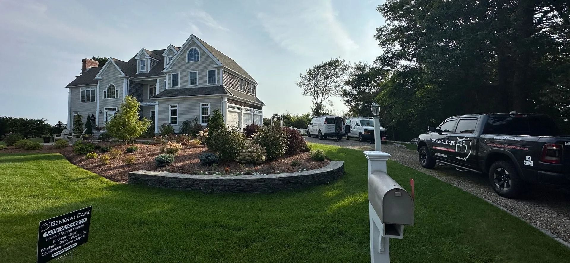 A large gray house with landscaping, a truck, and a mailbox on a sunny day.