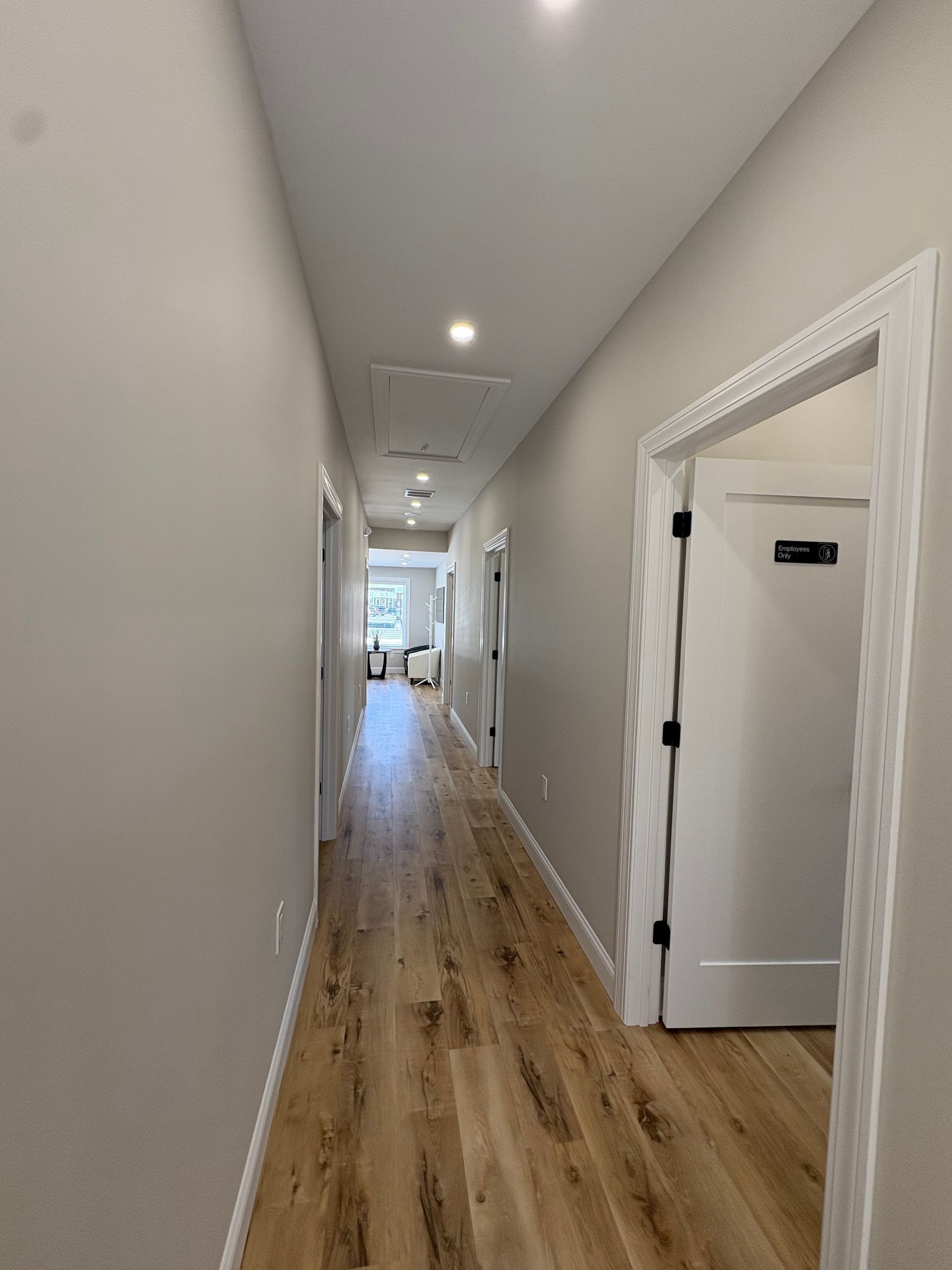Long hallway with light wood flooring, white trim, and doors. Recessed lighting in ceiling.