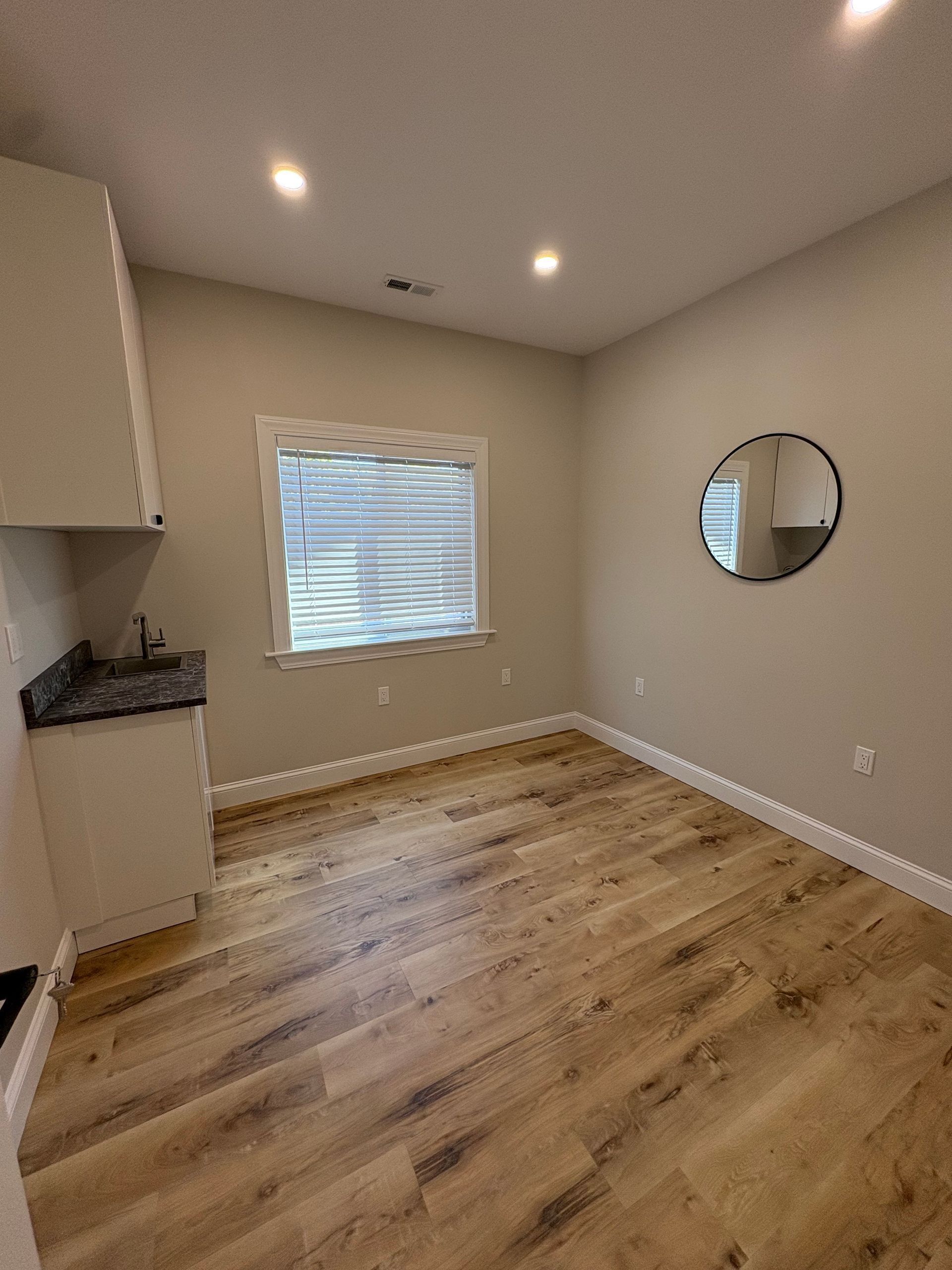 Empty room with wood-look flooring, a small cabinet with sink, window, and round mirror.