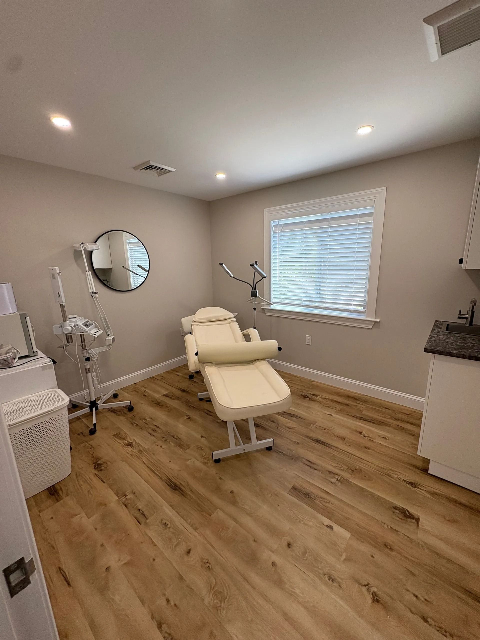 Aesthetician treatment room with beige chair, equipment, round mirror, and wood flooring.