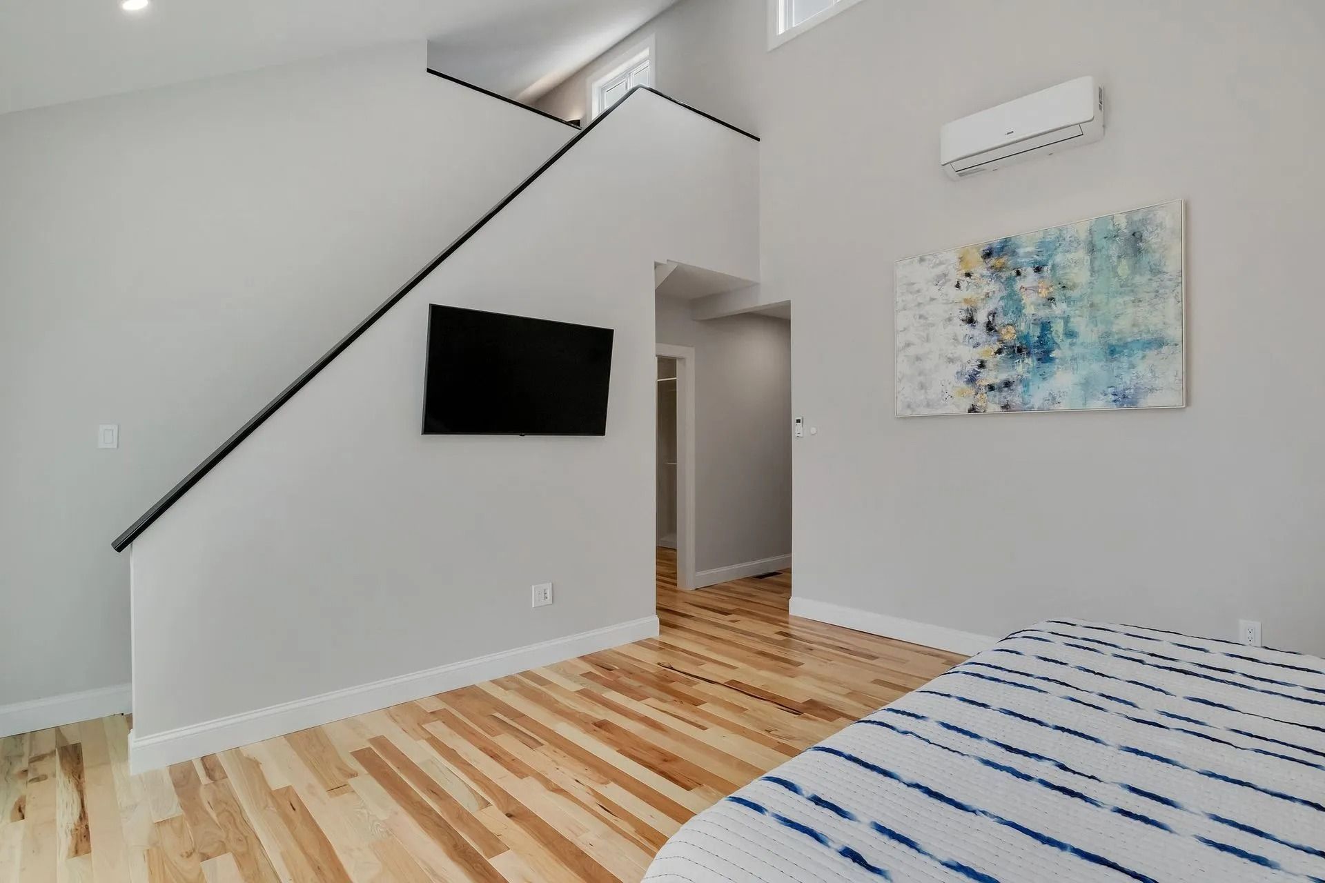Bedroom with wood floors, stairs, TV, art, and air conditioner on the wall. Light grey walls.