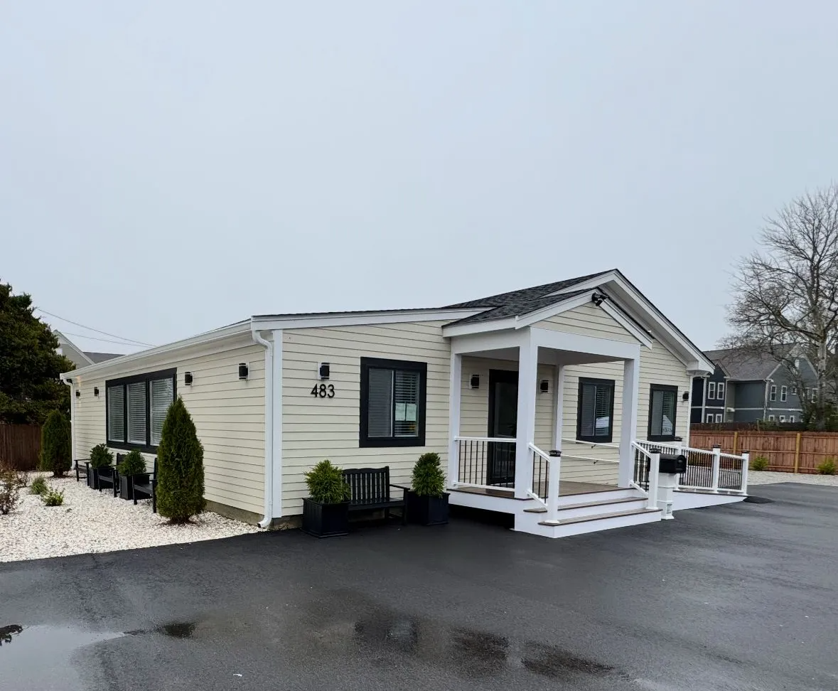 A light yellow, one-story building with black-framed windows and a small porch. The building sits on asphalt.