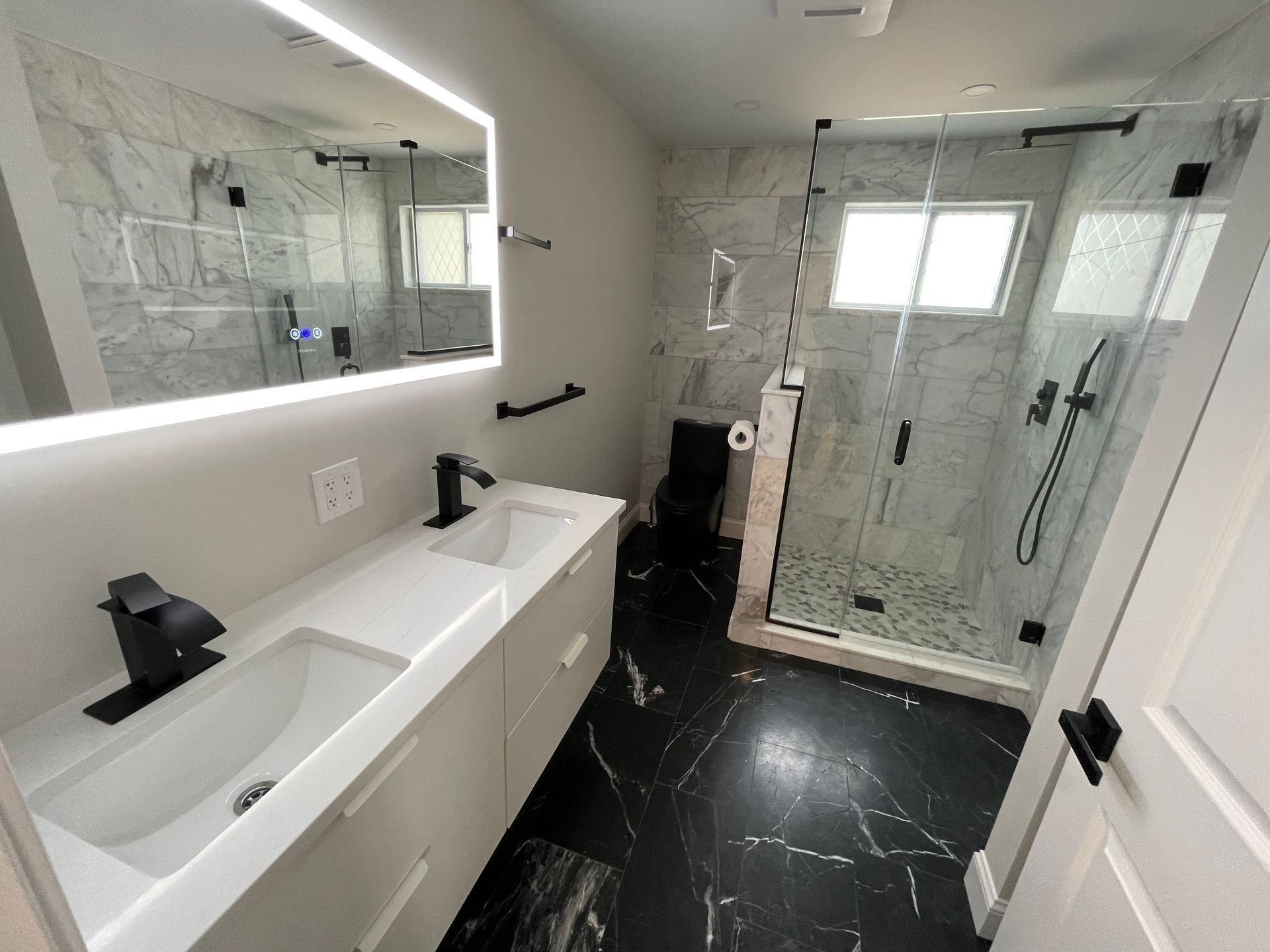 Modern bathroom with white countertops, black fixtures, marble tile walls, and a glass shower.