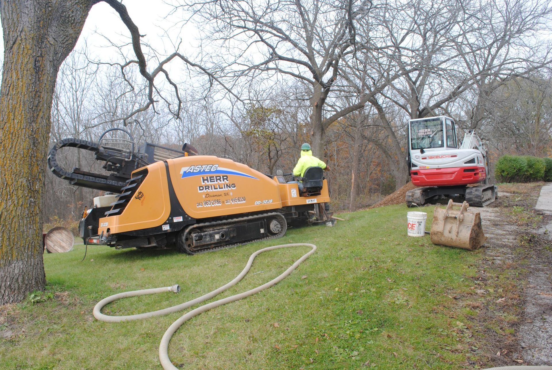 A horizontal directional drill and compact excavator on a grassy hillside. A worker operates the drill.