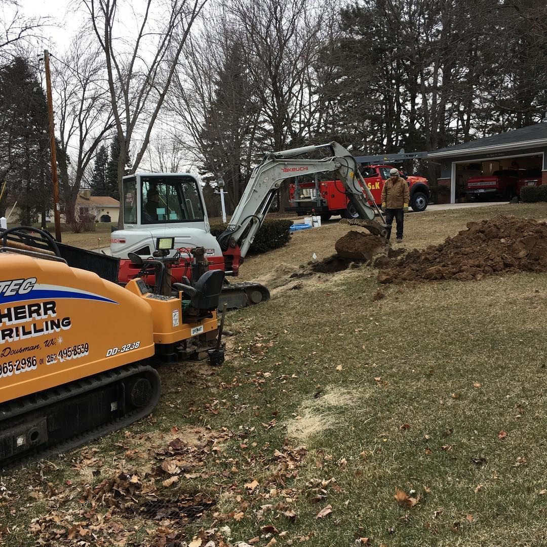 Construction site with drill, excavator, and man digging on a grassy area, residential background.