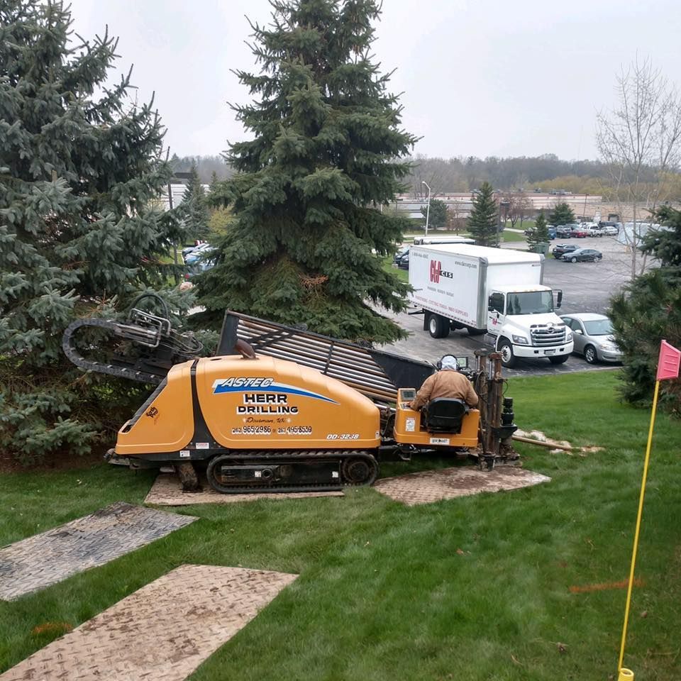 Yellow directional drilling machine in grassy area, person operating. White truck, trees, and cars in the background.