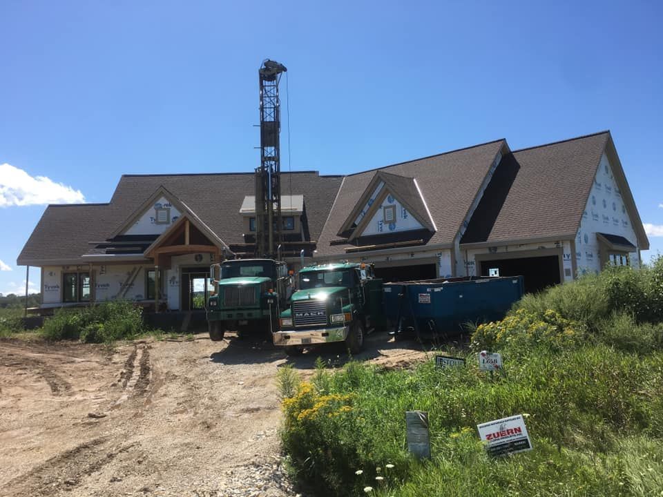 Trucks drilling a well in front of a partially constructed house on a sunny day.