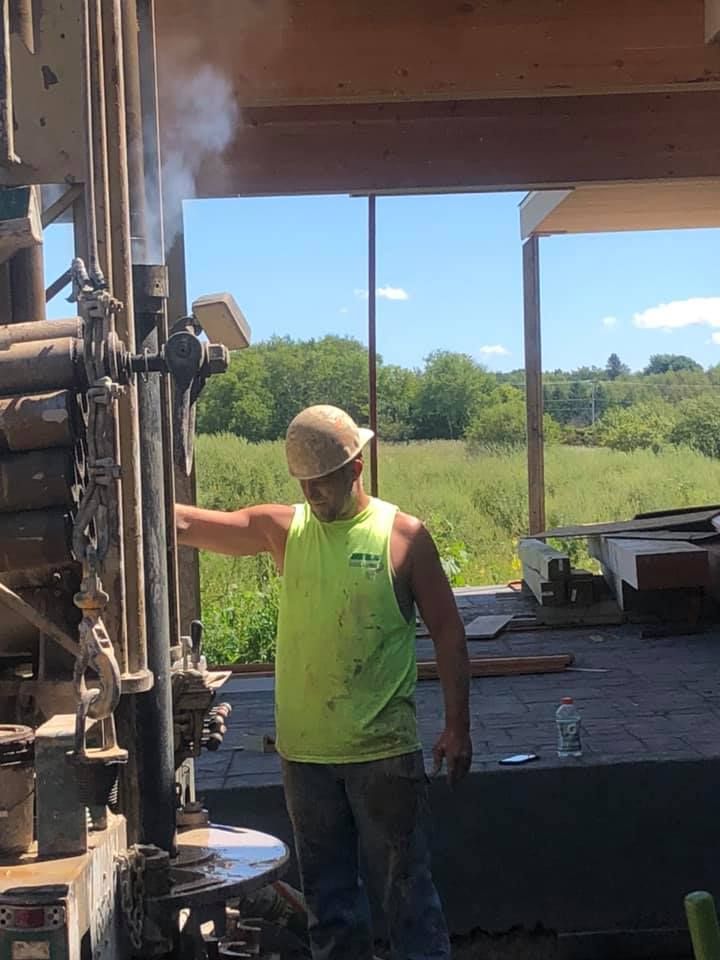 Man in safety vest operates drilling equipment outdoors. Steam rises, sunlight visible.