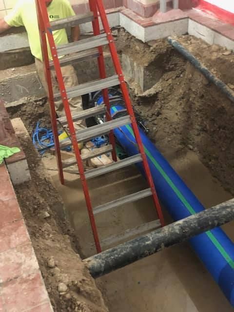 Construction site: worker near red ladder in trench, blue pipe visible.