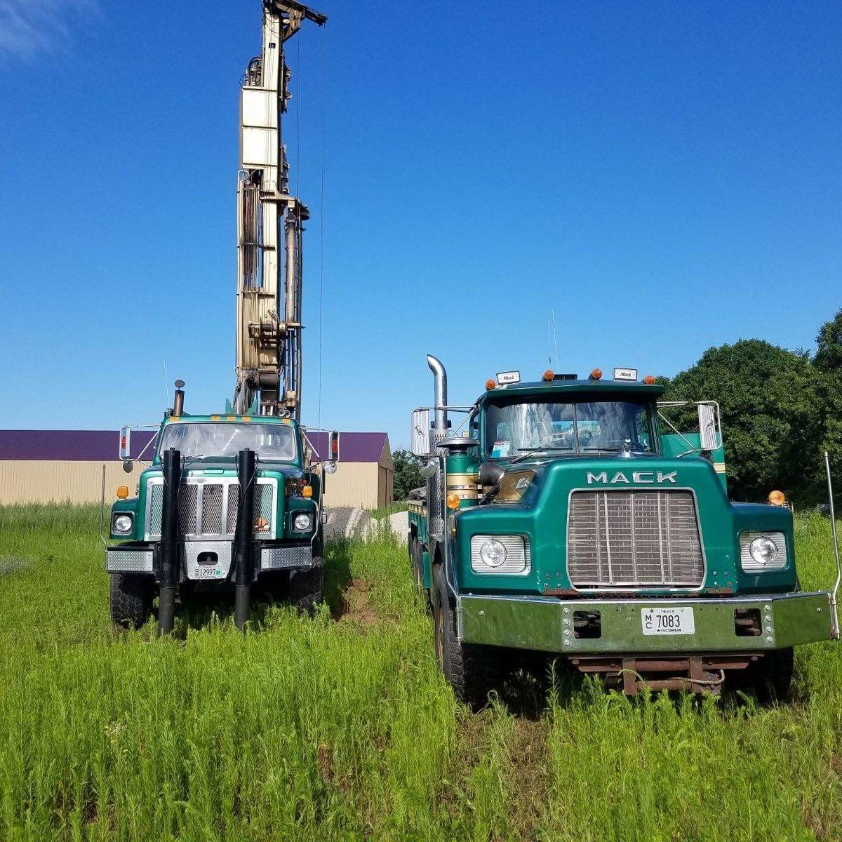 Two green Mack trucks in a grassy field; one with a drilling rig against a blue sky.