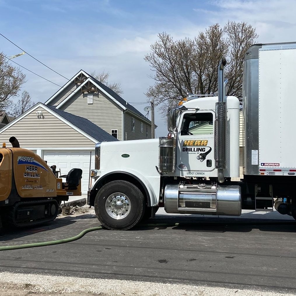 White semi-truck parked near a construction site. A small machine is visible to the left.  Gray house in the background.