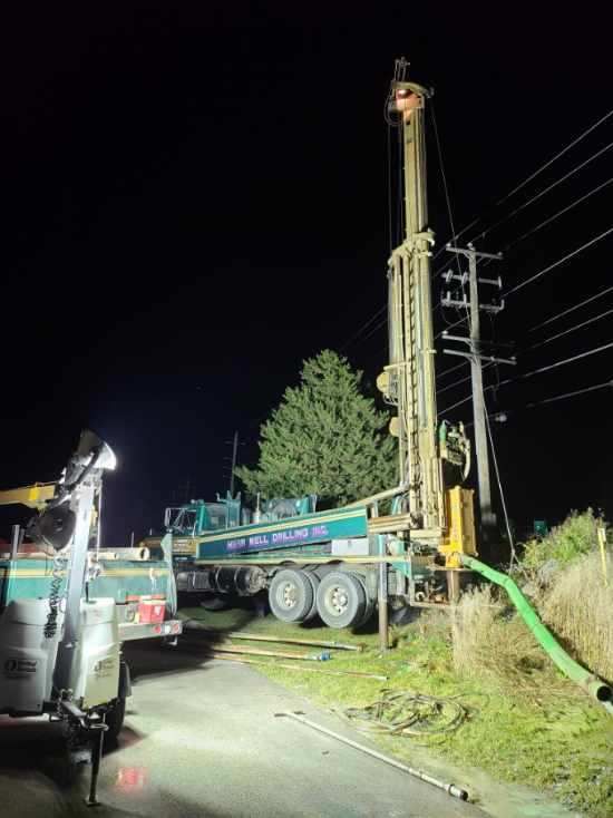Drilling rig next to a utility pole at night. Green and white truck, hose, and lights.