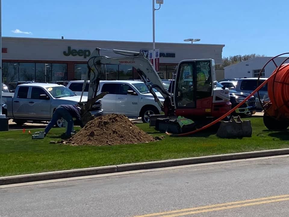 Construction workers installing fiber optic cable on a grassy area near a Jeep dealership.