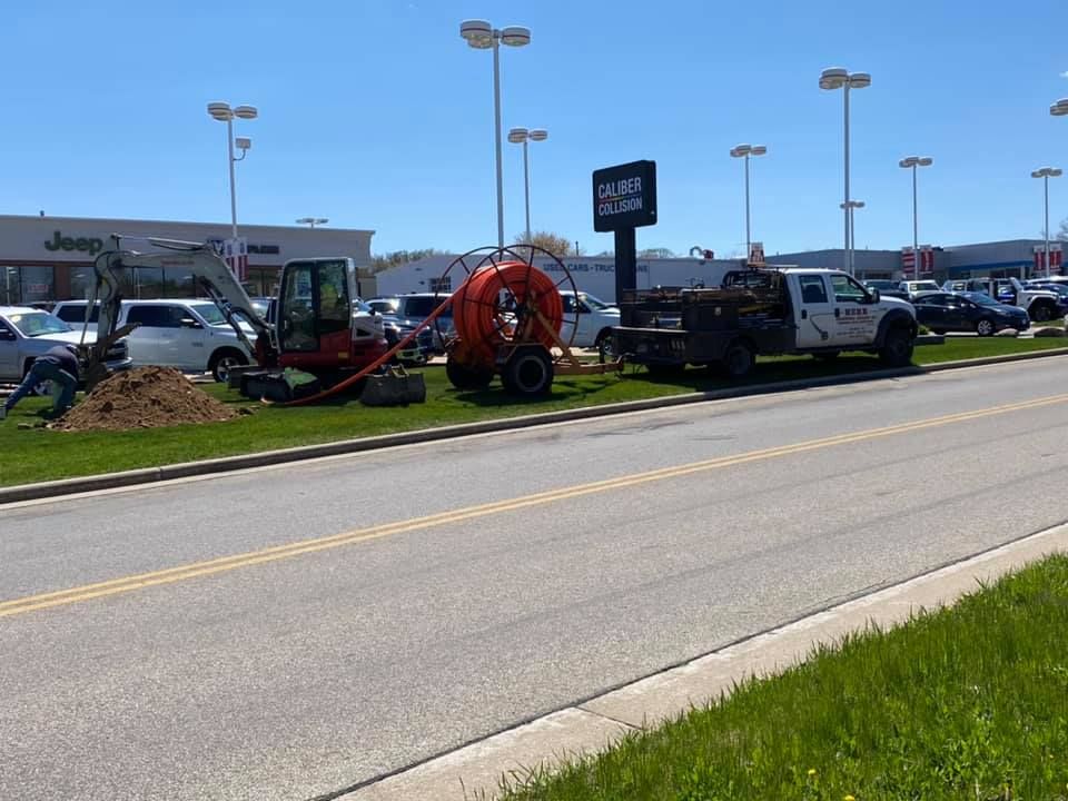 Construction equipment on a grassy median near a car dealership. Workers dig and operate machinery.