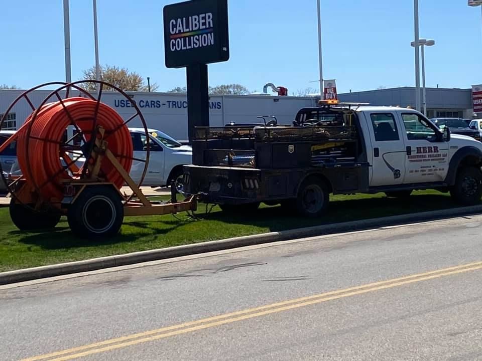 White truck with a cable reel trailer parked near a Caliber Collision sign.