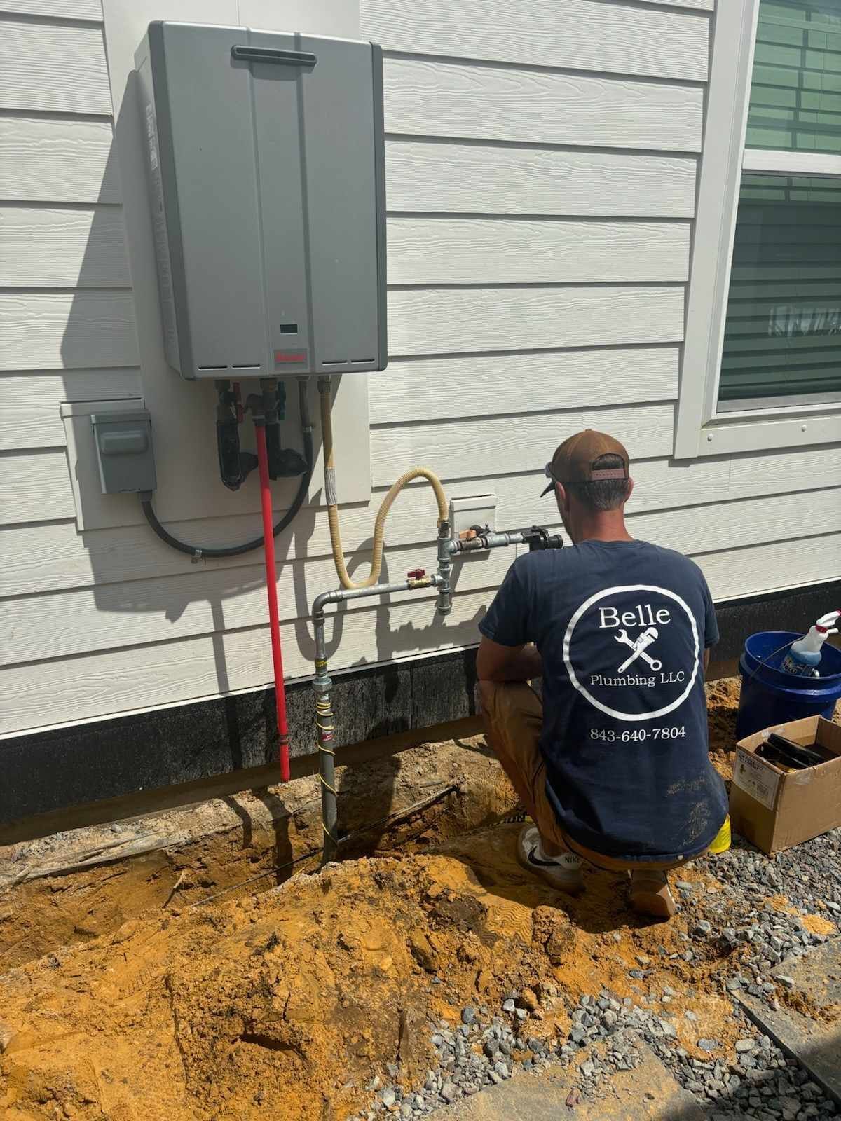 Worker kneeling in a dirt trench beside an electrical panel and utility piping on a house exterior