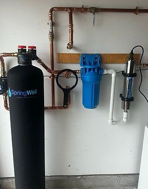 Water filtration system with black tank, blue filter housing, and clear inline filter mounted on a wall