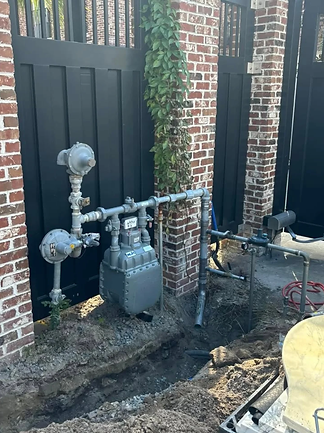 Gas meter and pipes mounted on a brick wall beside a black gate, with muddy ground below.