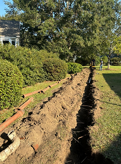 Long trench dug beside a hedge and lawn, with exposed pipes and a worker in the distance.