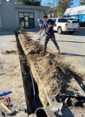 Worker digging a trench beside a building, with a utility line exposed and tools on the ground.