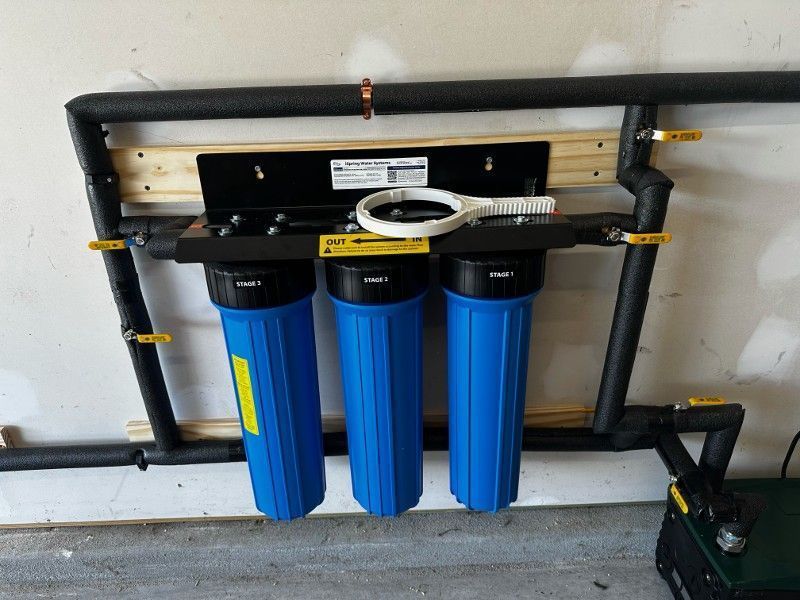 Wall-mounted blue water filtration system with three blue canisters and black piping in a utility room