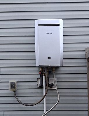 White Rinnai tankless water heater mounted on house siding with exposed pipes and hoses