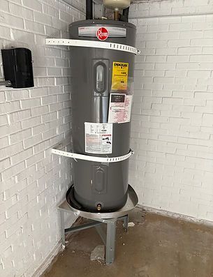 Gray water heater in a utility room against a white brick wall, with labels and a metal stand.