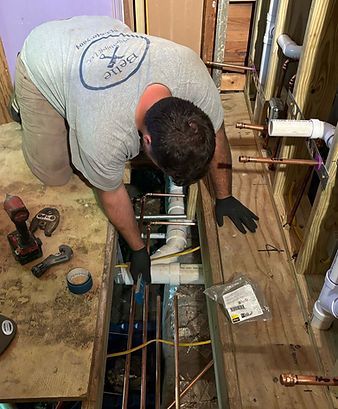 Worker kneeling over exposed floor pipes in a bathroom renovation, using tools beside scattered fittings.
