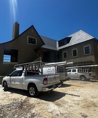 Two white pickup trucks parked in front of a large gray house under construction.
