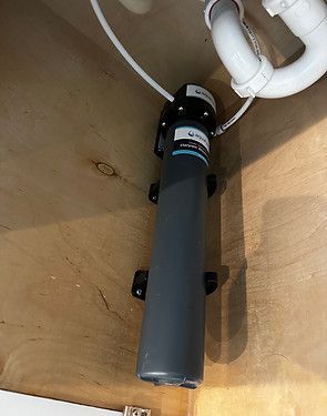 Gray cylindrical water filter mounted under a sink with white plumbing pipes above.